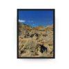 Vertical black-framed photograph of rugged Badlands rock formations at Dinosaur Provincial Park under a bright blue Alberta sky.